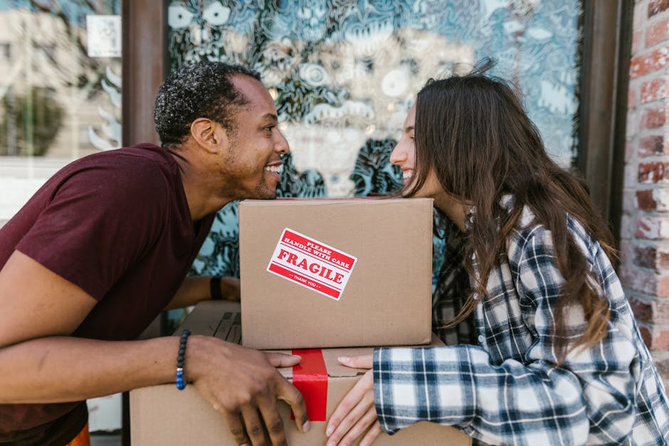 Two women in casual clothing are surrounded by multiple cardboard boxes, some sealed with red tape, in a residential setting with a textured grey wall in the background. One woman, wearing a checked s